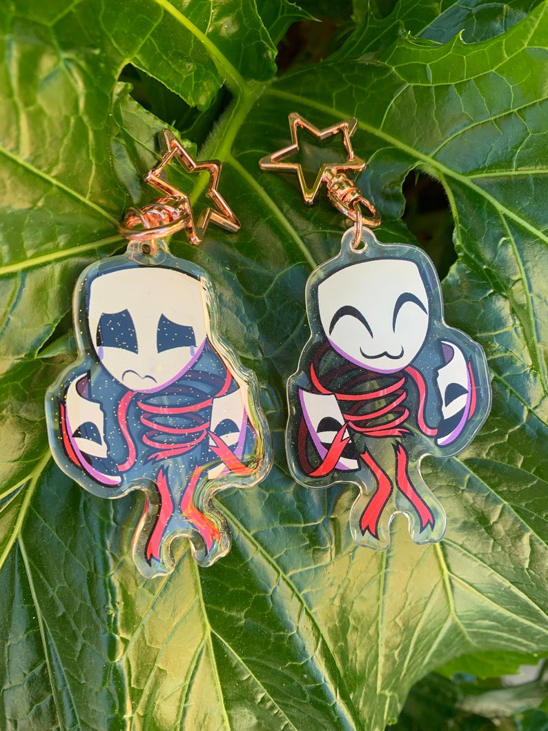 May include: Two clear acrylic keychains featuring a cartoon character with a red, white, and black mask. The character is holding a red ribbon with a white mask on it. The keychains are attached to gold star-shaped charms.