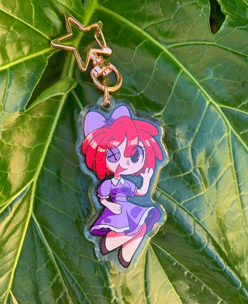 May include: A purple and red cartoon character keychain with a star-shaped charm. The character has red hair, a purple dress, and a white eye patch.
