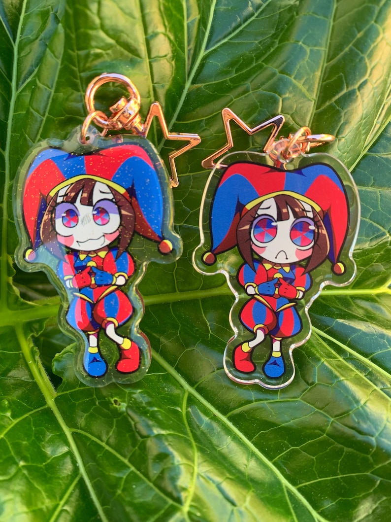 May include: Two clear acrylic keychains featuring a cartoon character dressed as a jester. The character has brown hair, red cheeks, and is wearing a red, blue, and yellow jester costume. The keychains are attached to gold star-shaped charms.