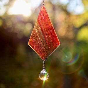 Könnte beinhalten: Ein roter und goldener rautenförmiger Glaswindspiel mit einem Kristallprisma, das daran hängt. Das Windspiel hängt an einer silbernen Kette.