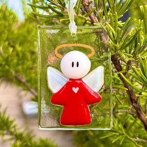Può includere: Un ornamento di vetro con un angelo bianco con un vestito rosso e un'aureola. L'angelo ha un piccolo cuore sul petto e ali con dettagli iridescenti. L'ornamento è appeso a un ramo.