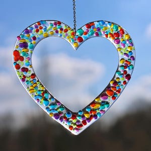 Könnte beinhalten: Ein farbenfroher gläserner Herz-Anhänger mit einer silbernen Kette, die oben hängt. Das Herz ist mit kleinen, bunten Glasstücken gefüllt.