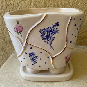 May include: A white ceramic planter with a kintsugi-style gold-lined cracked pattern. The surface features blue polka dots, blue floral illustrations, and small pink flower motifs. The planter sits on a matching white ceramic saucer base.