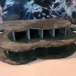 Könnte beinhalten: Dreistufige, dunkelgraue Schiefer-Aquariendekoration. Die Ebenen sind unregelmäßig geformt und werden von vertikalen Schieferstücken getragen, wodurch kleine höhlenartige Räume entstehen. Der Hintergrund zeigt eine verschwommene Wasserlandschaft.