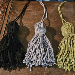 Könnte beinhalten: Drei dekorative Quasten in Schwarz, Grau und Grün. Die Quasten sind aus Garn gefertigt und haben einen langen, fließenden Fransen.