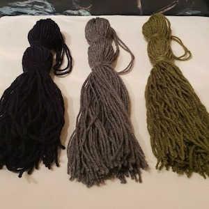 Könnte beinhalten: Drei Quasten aus Garn in einer Reihe. Die Quasten sind in den Farbtönen Schwarz, Grau und Olivgrün gehalten. Jede Quaste besteht aus dickem Garn mit einer geschlungenen Oberseite und einem langen, ausgefransten Boden. Die Quasten liegen auf einer weißen Oberfläche.