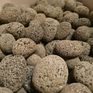 Könnte beinhalten: Nahaufnahme eines Haufens glatter, grauer Flusssteine. Die Steine sind unterschiedlich groß und geformt und weisen gesprenkelte Muster auf. Sie haben ein natürliches, verwittertes Aussehen mit einer neutralen Farbpalette aus Grau und Beige. Das Bild deutet auf natürliche Dekorationselemente hin.