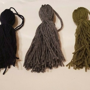 Könnte beinhalten: Drei Garnquasten in Schwarz, Grau und Grün. Die Quasten bestehen aus weichem, flauschigem Garn und haben einen langen, fließenden Fransen.