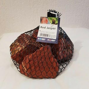 Könnte beinhalten: Ein schwarzer Netzbeutel gefüllt mit roten Jaspissteinen. Ein am Beutel befestigtes Etikett trägt die Aufschrift "Red Jasper" und zeigt einen Barcode sowie das "Hobby"-Logo. Die Steine sind tiefrotbraun und variieren in Größe und Form.