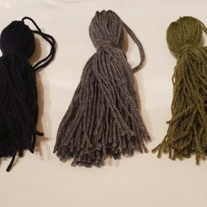 Könnte beinhalten: Drei Garnquasten in Schwarz, Grau und Grün. Die Quasten bestehen aus weichem, flauschigem Garn und haben einen langen, fließenden Fransen.