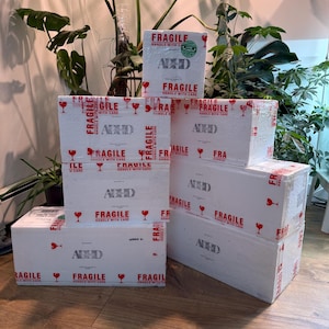 May include: A stack of white cardboard boxes with red "Fragile" and "Handle with Care" text and wine glass icons. The boxes also feature the word "ADHD" in a stylized font. The boxes are stacked on a wooden floor.