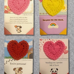 May include: Four greeting cards with crocheted heart accents. Each card features a different colored heart: pink, yellow, red, and another pink. The cards have various messages and illustrations, including a cow, a dog, a panda, and a rainbow.