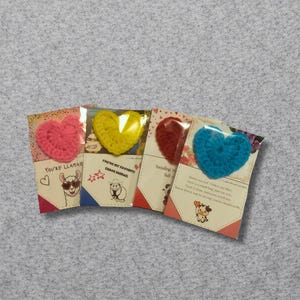 May include: Four greeting cards with crochet heart embellishments. The hearts are pink, yellow, red, and blue. Each card has a different design and text, including a llama, sheep, and cow illustration.