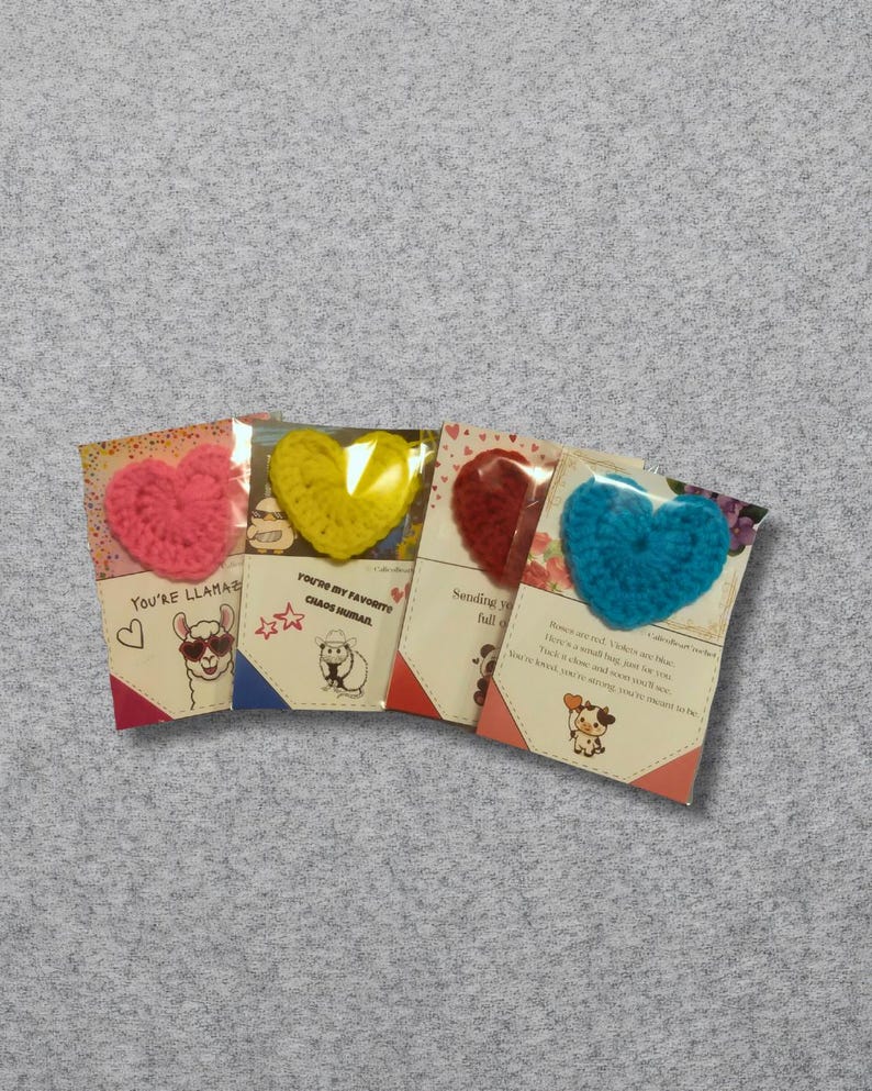 May include: Four greeting cards with crochet heart embellishments. The hearts are pink, yellow, red, and blue. Each card has a different design and text, including a llama, sheep, and cow illustration.