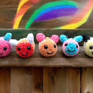 May include: Five crocheted plush toys in the shape of smiling cartoon characters. The toys are pink, orange, red, blue, and yellow. They are lined up on a wooden surface.