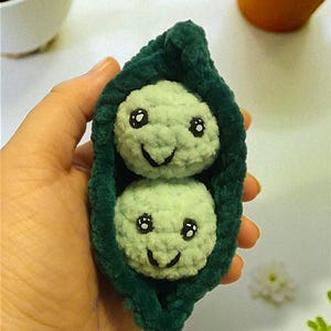 May include: A handmade crochet pea pod with two smiling pea characters. The pea pod is dark green, and the peas are light green with black eyes and smiling faces. The item is held in a hand.