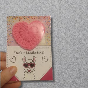 May include: A pink crocheted heart attached to a card that says "You're Llamazing!" with a cartoon llama wearing heart-shaped sunglasses. The card has a dotted background and two heart illustrations.