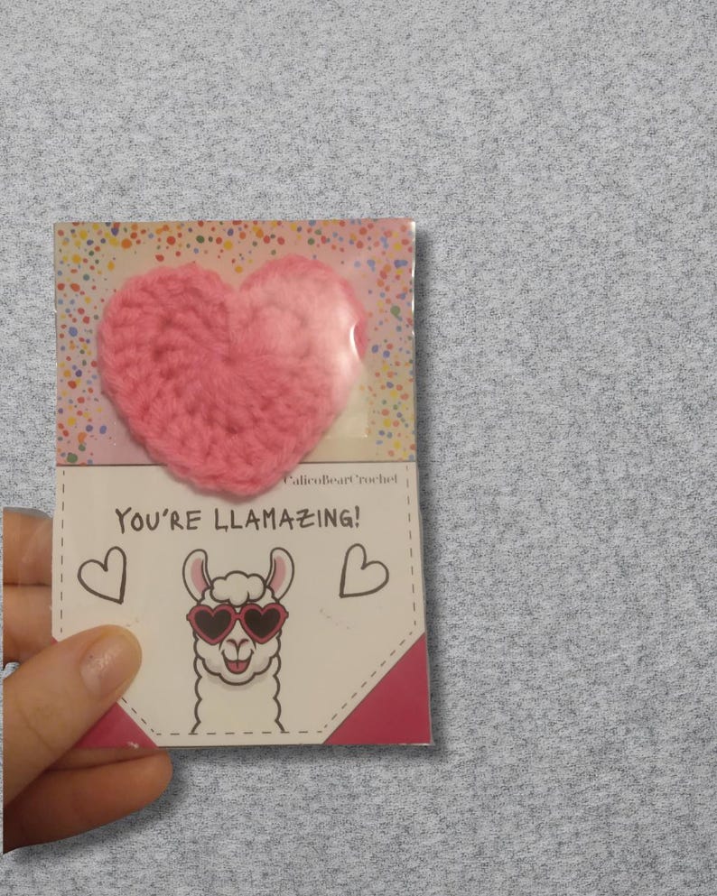 May include: A pink crocheted heart attached to a card that says "You're Llamazing!" with a cartoon llama wearing heart-shaped sunglasses. The card has a dotted background and two heart illustrations.
