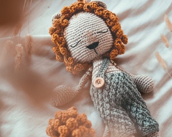 Peut inclure: Un jouet en crochet en forme de lion avec une crinière brune et une tenue grise et verte. Le jouet est couché sur un fond en tissu blanc.