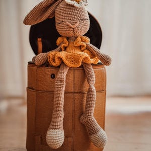 Puede incluir: Una muñeca de conejito de ganchillo hecha a mano con patas largas y orejas caídas, que lleva un vestido amarillo y una corona de flores. El conejito está sentado en un contenedor redondo de cuero marrón. La paleta de colores de la muñeca incluye beige, amarillo y marrón.