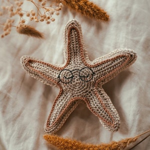May include: A crocheted starfish with a brown and beige color scheme. The starfish has black eyes that resemble glasses.