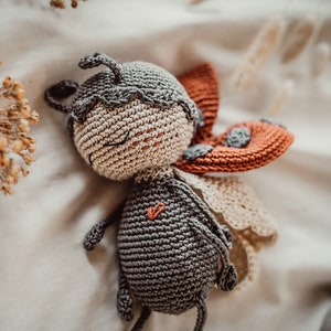 May include: A crocheted ladybug toy with a grey body, beige face, and orange wings with white accents. The toy has a heart-shaped design on its chest.