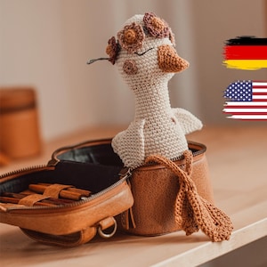 May include: A handmade crochet goose doll with a floral crown, resting inside a brown leather bag. The goose has a beige body, brown beak, and closed eyes. The bag is open, revealing a glimpse of its contents.