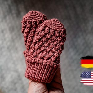 Puede incluir: Un par de mitones de crochet rosa con un patrón texturizado. Los mitones se sostienen en una mano, con las puntas de los dedos de los mitones visibles.