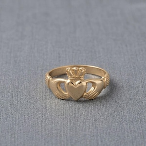 Puede incluir: Un anillo Claddagh dorado con un diseño de corazón, corona y manos. El corazón está en el centro, coronado y flanqueado por manos. El anillo está sobre un fondo gris.