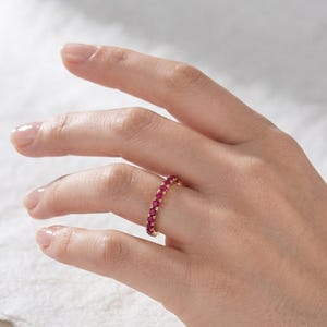 May include: A gold eternity band ring featuring a continuous row of small, round, vibrant pinkish-red gemstones set in a shared prong setting. The ring encircles the finger, showcasing a uniform design with polished metal accents between each stone.