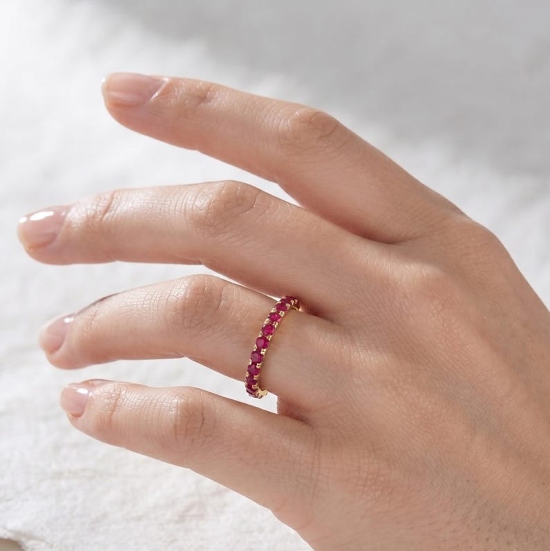 May include: A gold eternity band ring featuring a continuous row of small, round, vibrant pinkish-red gemstones set in a shared prong setting. The ring encircles the finger, showcasing a uniform design with polished metal accents between each stone.