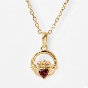 May include: A gold-colored Claddagh pendant necklace with a red heart-shaped gemstone. The pendant features a heart, crown, and hands design, suspended from a delicate chain. The necklace is displayed against a white background.