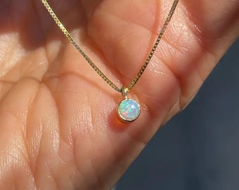 May include: A delicate gold necklace featuring a small, round opal pendant. The opal displays a range of colors, including white, blue, and green. The chain is a fine, square-linked design.