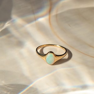 May include: A gold ring featuring an oval, pale opal gemstone. The ring is set on a light-toned surface, with shadows and light reflections. The ring is a piece of jewellery.
