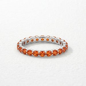 May include: A silver eternity band ring featuring a continuous row of round-cut, vibrant orange gemstones set in a polished metal frame. The circular design showcases uniform stones that wrap around the entire circumference of the band.