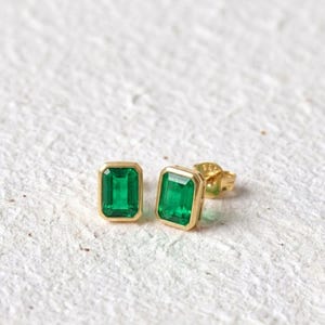 May include: A pair of stud earrings featuring rectangular emerald-cut green gemstones set in gold bezels. Each earring has a polished gold post and a matching gold butterfly clutch backing, showcasing a clean, geometric design with vibrant green hues.