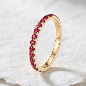 May include: A gold band ring featuring a continuous row of small, round, vibrant red rubies set in a shared prong setting. The metal has a polished finish, and the stones are arranged along the outer circumference of the circular band.