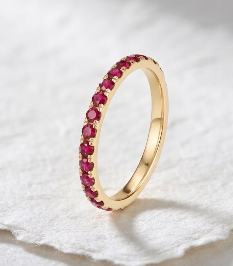 May include: A gold band ring featuring a continuous row of small, round, vibrant red rubies set in a shared prong setting. The metal has a polished finish, and the stones are arranged along the outer circumference of the circular band.