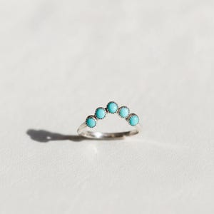May include: A silver ring with five turquoise stones, each approximately 3 mm in diameter, arranged in an arc. The ring sits on a white surface, with a shadow to the left.