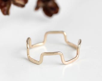 Pode incluir: Um delicado anel de ouro com um design geométrico único. O anel apresenta uma série de formas angulares conectadas, criando uma estética moderna e minimalista. O anel está sobre um fundo branco macio, com um galho desfocado ao fundo.