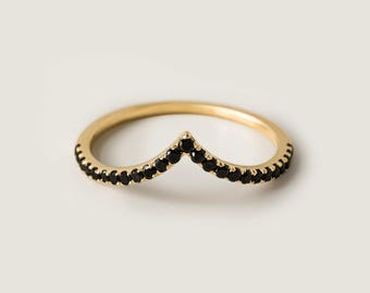 May include: A gold-colored ring with a chevron design. The ring is adorned with small, black gemstones along the chevron shape. The ring is set against a plain, off-white background.