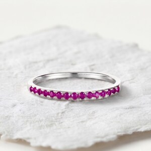 May include: A thin silver-toned metal band ring featuring a row of small, round, vibrant magenta-pink gemstones set along the front half of the band. The ring has a polished finish and a simple, slender design with a consistent width throughout.