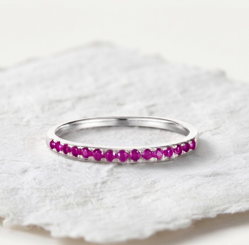 May include: A thin silver-toned metal band ring featuring a row of small, round, vibrant magenta-pink gemstones set along the front half of the band. The ring has a polished finish and a simple, slender design with a consistent width throughout.
