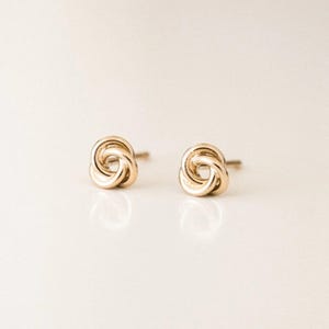 May include: A pair of gold-coloured stud earrings. Each earring features a knot design, with the metal looped and intertwined to create the knot shape. The earrings are displayed on a white surface.