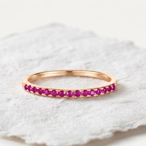 May include: A thin rose gold band ring featuring a row of small, round, vibrant magenta-pink gemstones set along the front half of the band. The metal has a smooth, polished finish, and the stones are uniform in size and color, creating a continuous line.