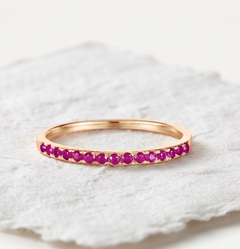 May include: A thin rose gold band ring featuring a row of small, round, vibrant magenta-pink gemstones set along the front half of the band. The metal has a smooth, polished finish, and the stones are uniform in size and color, creating a continuous line.