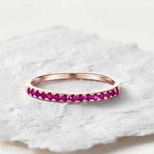 May include: A thin rose gold band ring featuring a continuous row of small, round, vibrant pinkish-red rubies set along the front half of the metal band. The ring has a polished finish and a delicate, slender profile with a consistent circular shape.