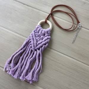 Puede incluir: Un collar de macramé morado con un anillo de madera y un cordón de gamuza marrón. El collar tiene una franja de borlas.