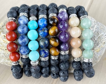 Puede incluir: Siete pulseras de roca de lava con cuentas de piedras preciosas de diferentes colores. Las pulseras están dispuestas en fila, con las cuentas rojas a la izquierda y las cuentas verdes a la derecha. Las cuentas son todas redondas y tienen un acabado liso.