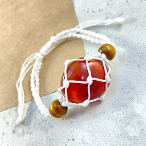 May include: A white cord bracelet with a red and orange gemstone encased in a white macrame net. The bracelet has two brown wooden beads.